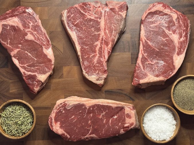 Four raw dry-aged steaks on a wood cutting board with salt, pepper, and rosemary
